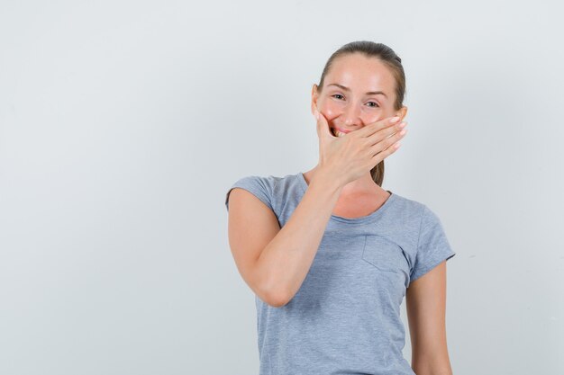 Jong wijfje die hand op mond in grijs t-shirt houden en vreugdevol, vooraanzicht kijken.