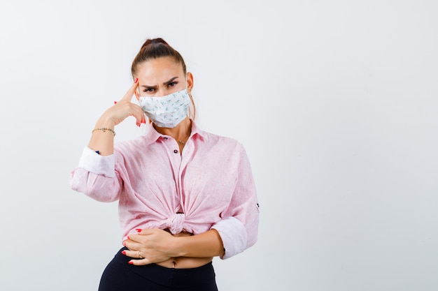 Jong wijfje in overhemd, broek, medisch masker dat zich in het denken stelt stelt en peinzend kijkt, vooraanzicht.