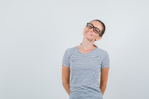 Jong wijfje in t-shirt, glazen die handen op rug houden en schattig, vooraanzicht kijken.