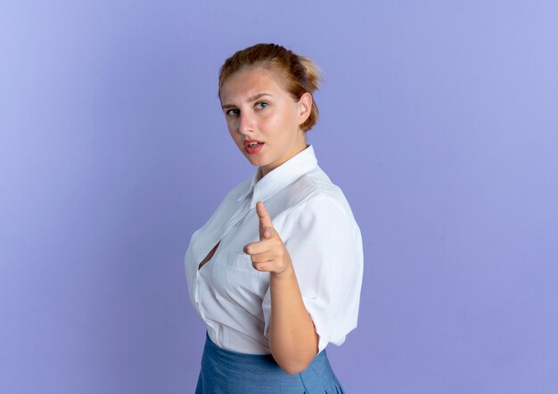 Jong zelfverzekerd blond Russisch meisje staat zijwaarts wijzend op camera geïsoleerd op paarse achtergrond met kopie ruimte