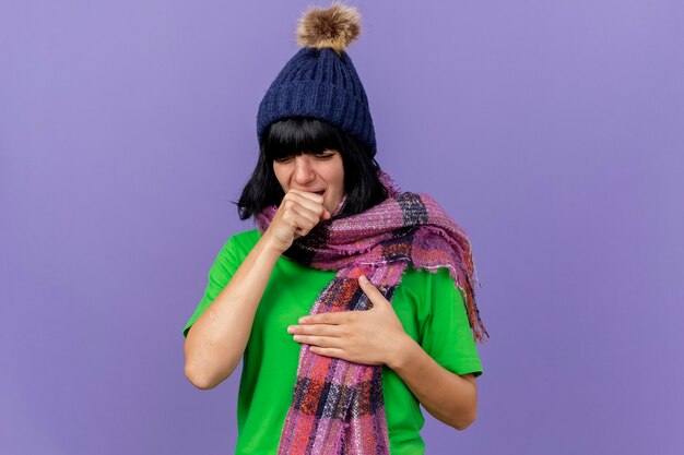 Jong ziek Kaukasisch meisje die de winterhoed en sjaal dragen die borst hoesten geïsoleerd op purpere achtergrond met exemplaarruimte aanraken
