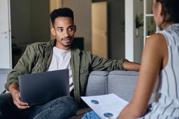 Jonge aantrekkelijke Afro-Amerikaanse man met laptop die samenwerkt met collega in moderne co-working space