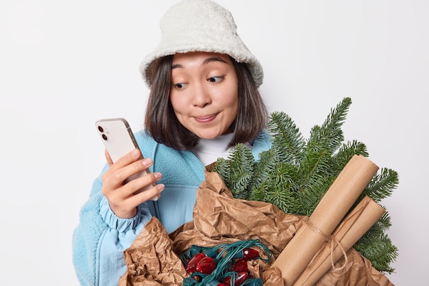 Jonge aantrekkelijke aziatische vrouw gekleed in winterkleren gebruikt mobiele telefoon om online te chatten draagt groene sparren takken gewikkeld in papieren slinger stuurt vrienden uitnodigingen voor nieuwjaarsfeest