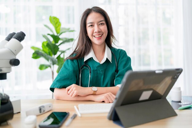 Jonge aantrekkelijke Aziatische vrouwelijke arts diagnose praten en kijken naar camera in videoconferentie Positieve arts zwaaien en online consultatie op digitale tablet laptop