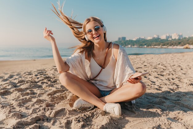 Jonge aantrekkelijke blonde lachende vrouw zittend op het strand in koptelefoon luisteren naar muziek in positieve stemming