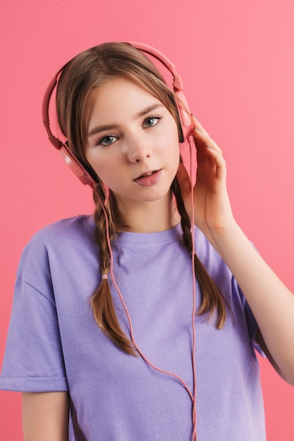 Jonge aantrekkelijke dame met twee vlechten in lila t-shirt luisteren muziek in koptelefoon bedachtzaam in de camera kijken terwijl tijd doorbrengen over roze achtergrond