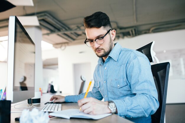 Jonge aantrekkelijke donkerharige man in glassess werkt met een computer en schrijven in notitieblok in kantoor. Hij draagt een blauw shirt, baard.