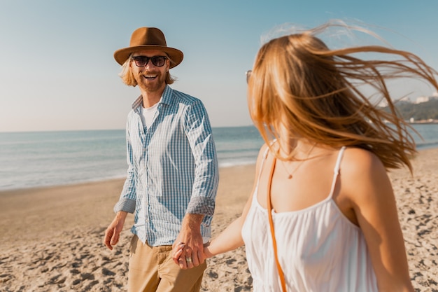 Gratis foto jonge aantrekkelijke glimlachende gelukkig man in hoed en blonde vrouw in witte jurk die samen op strand loopt