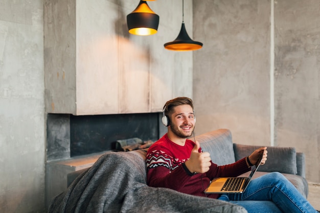 Jonge aantrekkelijke glimlachende man om thuis te zitten in de winter, koptelefoon luisteren, student online studeren, rode gebreide trui dragen, vasthouden aan laptop, freelancer, duim opdagen, positief teken