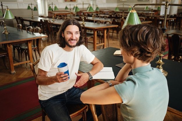 Jonge aantrekkelijke glimlachende mannelijke student die gelukkig met vriend praat tijdens studiepauze in bibliotheek van universiteit