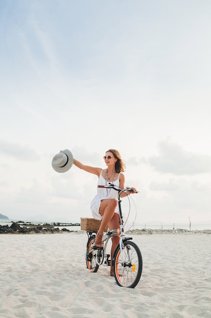 Jonge aantrekkelijke glimlachende vrouw in witte kleding die op tropisch strand op fiets berijdt die hoed en zonnebril draagt