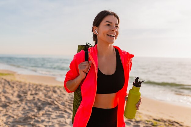 Jonge aantrekkelijke lachende vrouw doet sport in de ochtend zonsopgang op zee strand yoga mat en fles water te houden, gezonde levensstijl, luisteren naar muziek op oortelefoons, roze windjack dragen