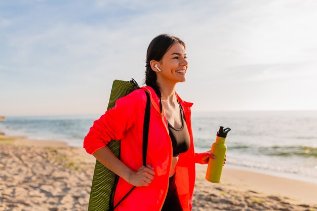 Jonge aantrekkelijke lachende vrouw doet sport in de ochtend zonsopgang op zee strand yoga mat en fles water te houden, gezonde levensstijl, luisteren naar muziek op oortelefoons, roze windjack dragen