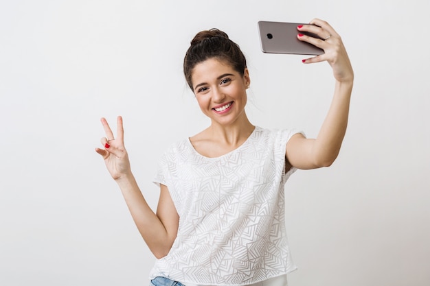 Jonge aantrekkelijke lachende vrouw in witte blouse met slimme telefoon, vredesteken, positieve emotie tonen, selfie foto maken,