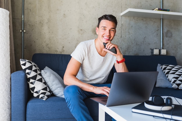 Jonge aantrekkelijke man zittend op de bank thuis werken op laptop online, via internet