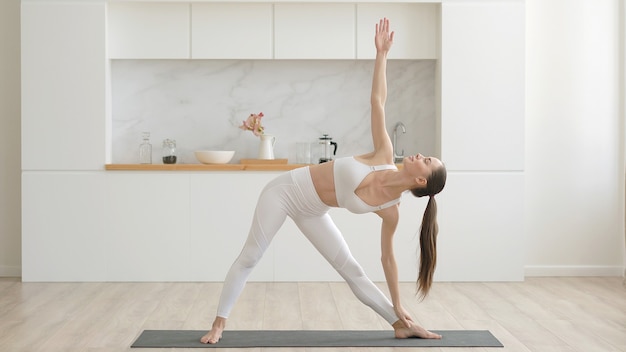 Jonge aantrekkelijke multi-etnische vrouw, gekleed in witte sportkleding broek en top doen stretching yoga-oefeningen op fitness mat in woonkamer gezondheidszorg en oefen mentale therapie thuis concept