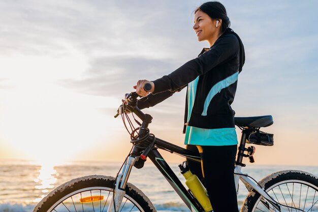 Jonge aantrekkelijke slanke vrouw fietsten, sport in de zomerstrand van de ochtendzonsopgang in sportfitnesskleding, actieve gezonde levensstijl, glimlachend gelukkig met plezier