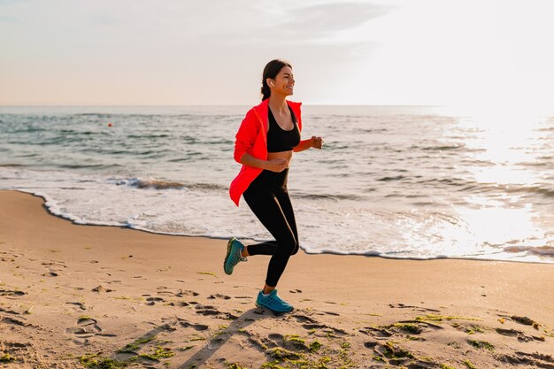 Jonge aantrekkelijke slanke vrouw sport beoefening in ochtend zonsopgang joggen op zee strand in sportkleding, gezonde levensstijl, luisteren naar muziek op oortelefoons, roze windjack dragen