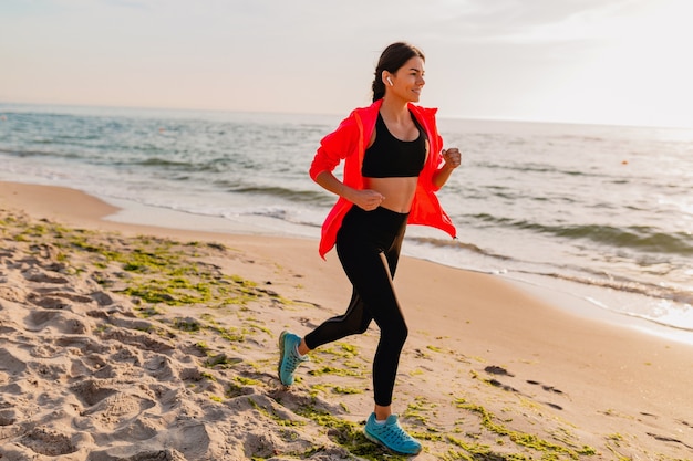 Jonge aantrekkelijke slanke vrouw sport beoefening in ochtend zonsopgang joggen op zee strand in sportkleding, gezonde levensstijl, luisteren naar muziek op oortelefoons, roze windjack dragen