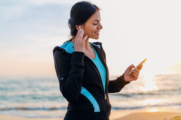 Jonge aantrekkelijke slanke vrouw sport beoefening op ochtend zonsopgang strand in sportkleding, gezonde levensstijl, luisteren naar muziek op draadloze oortelefoons met smartphone, glimlachend gelukkig plezier