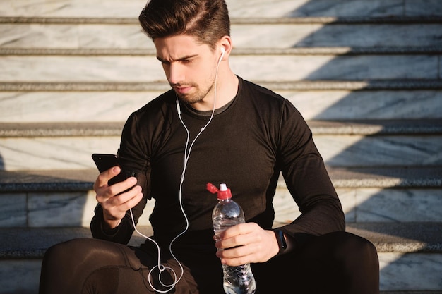 Jonge aantrekkelijke sportieve man in oortelefoons die zorgvuldig mobiele telefoon op trappen gebruikt na training buiten