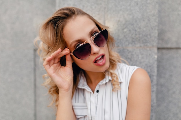 Jonge aantrekkelijke stijlvolle blonde vrouw wandelen in de stad straat in zomer mode stijl jurk dragen van een zonnebril, lachen verrast