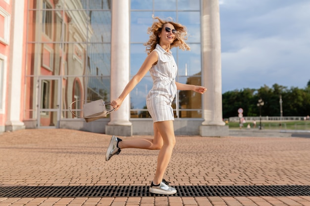 Jonge aantrekkelijke stijlvolle vrouw springen grappig uitgevoerd in sneakers in stad straat in zomer mode stijl witte jurk dragen van een zonnebril en handtas