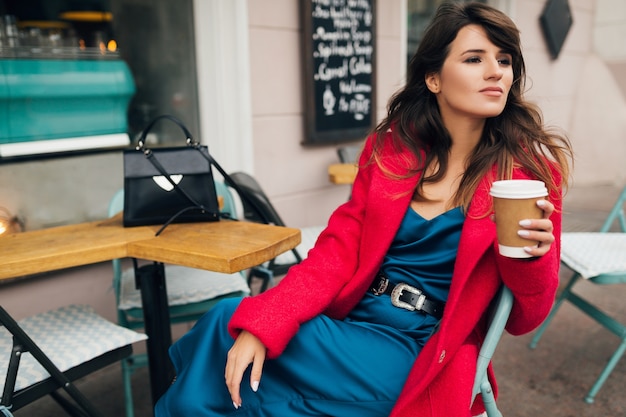 Jonge aantrekkelijke stijlvolle vrouw zitten in stad straat café in rode jas koffie drinken dragen blauwe jurk