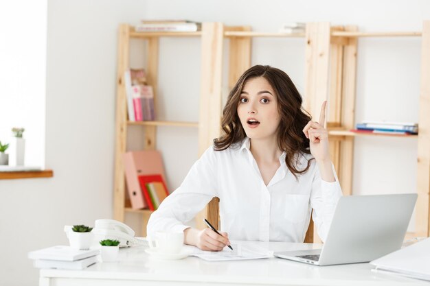 Jonge aantrekkelijke vrouw bij een modern bureau dat met laptop werkt en aan iets denkt