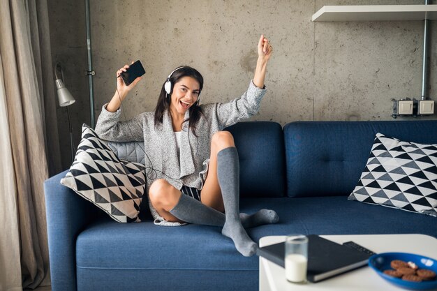 Jonge aantrekkelijke vrouw in casual outfit ontspannen thuis, vrije tijd, plezier hebben, glimlachend luisteren naar muziek op koptelefoon, dansen zittend op de bank, vrolijk, blij, kousen dragen, trui