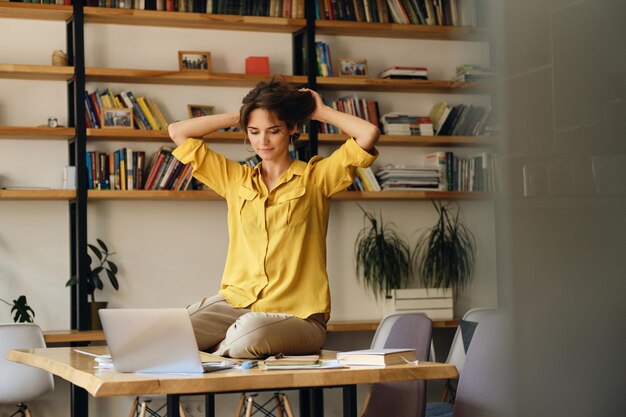 Jonge aantrekkelijke vrouw in geel shirt zittend op een bureau terwijl ze dromerig aan het werk is op een laptop in een modern kantoor