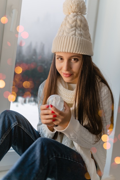 Jonge aantrekkelijke vrouw in stijlvolle witte gebreide trui, sjaal en muts om thuis te zitten op de vensterbank met Kerstmis glas sneeuw bal aanwezig decoratie, winter boszicht, lichten bokeh houden