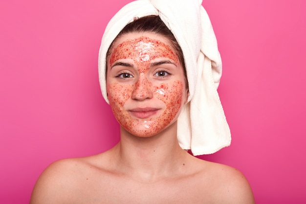 Gratis foto jonge aantrekkelijke vrouw met witte handdoek op haar hoofd, heeft naakte lichaam, smilling geïsoleerd over roze muur in studio, kijkt direct naar de camera, met rode scrub op haar gezicht.