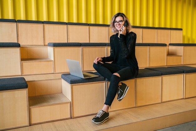 Jonge aantrekkelijke vrouw zitten in collegezaal, bezig met laptop, bril, moderne auditorium, student onderwijs online, freelancer, glimlachen, praten aan de telefoon