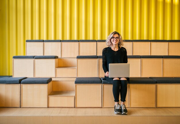 Jonge aantrekkelijke vrouw zitten in de collegezaal, die op laptop werkt, een bril draagt, een modern auditorium, online studentenstudie, freelancer, glimlachen, tieneropstarten