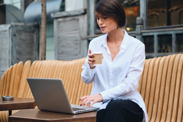 Jonge aantrekkelijke zakenvrouw die op laptop werkt tijdens de koffiepauze in café op straat