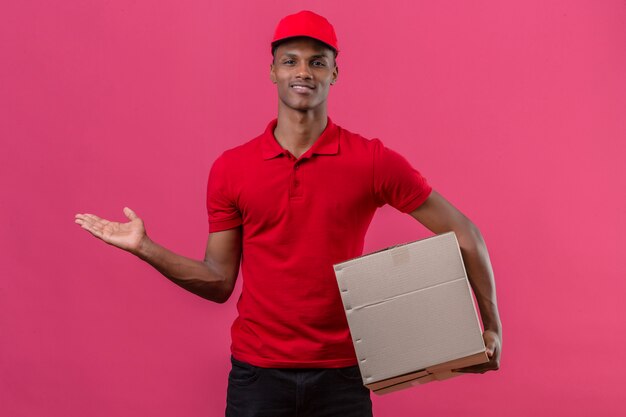 Jonge Afrikaanse Amerikaanse leveringsmens die rood poloshirt draagt en kartondoos houdt en iets voorstelt dat hand aan de kant over geïsoleerd roze richt