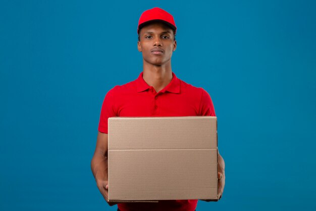 Jonge Afrikaanse Amerikaanse leveringsmens die rood poloshirt en GLB-de doos dragen die van het holdingskarton zich met ernstig gezicht over geïsoleerd blauw bevinden