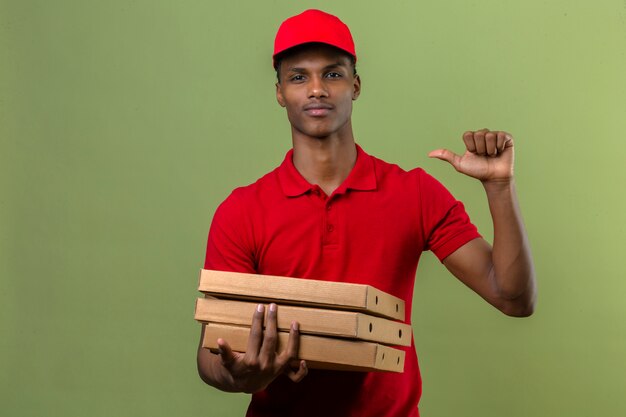 Jonge Afrikaanse Amerikaanse leveringsmens rood poloshirt dragen en GLB die zich met stapel pizzadozen bevinden die vinger wijzen aan zich het zekere kijken over geïsoleerde groen