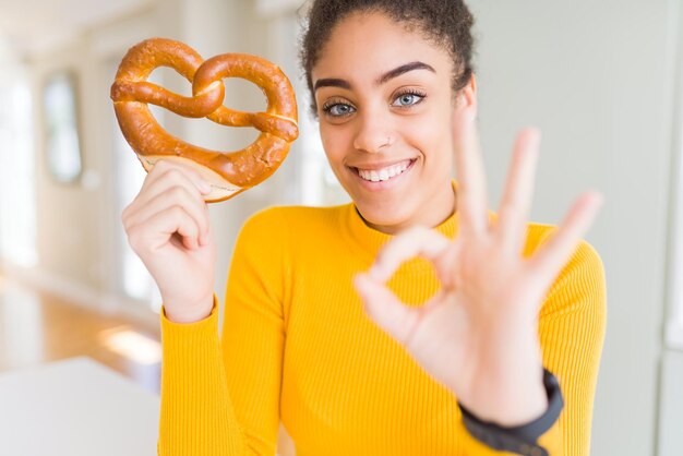 Gratis foto jonge afrikaanse amerikaanse vrouw die duitse zoute gebakjepretzel eet die ok teken met vingers uitstekend symbool doet