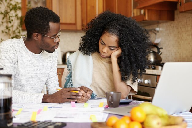 Jonge Afrikaanse familie die financiële crisis onder ogen ziet. Man met bril probeert zijn mooie vrouw te kalmeren, houdt haar hand vast en vertelt dat alles goed komt terwijl hij de financiën in de keuken beheert