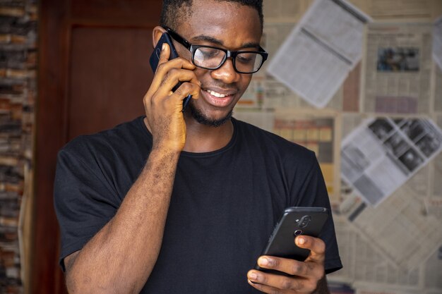 Jonge Afrikaanse man met een bril die aan de telefoon praat terwijl hij een andere in een kamer gebruikt