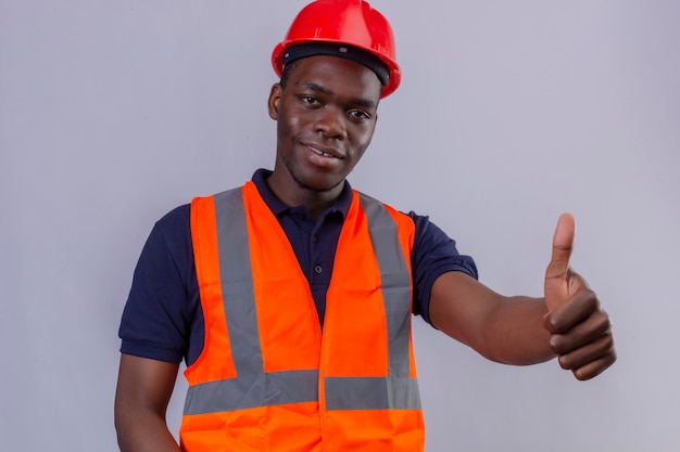 Jonge Afro-Amerikaanse bouwersmens die bouwvest en veiligheidshelm met glimlach op gezicht draagt ?? die duim omhoog toont