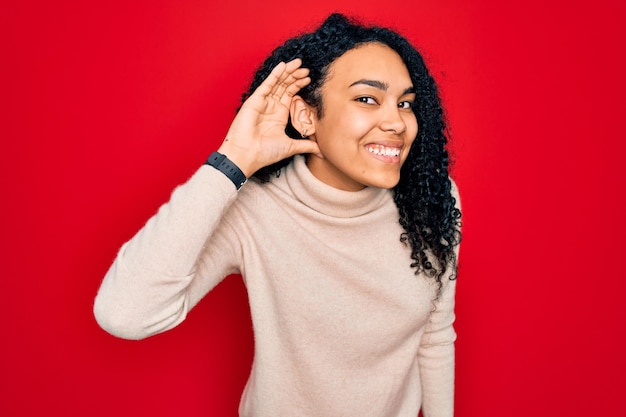 Jonge afro-amerikaanse gekrulde vrouw, gekleed in casual coltrui over rode achtergrond glimlachend met hand over oor luisterend naar geruchten of roddels Doofheid concept