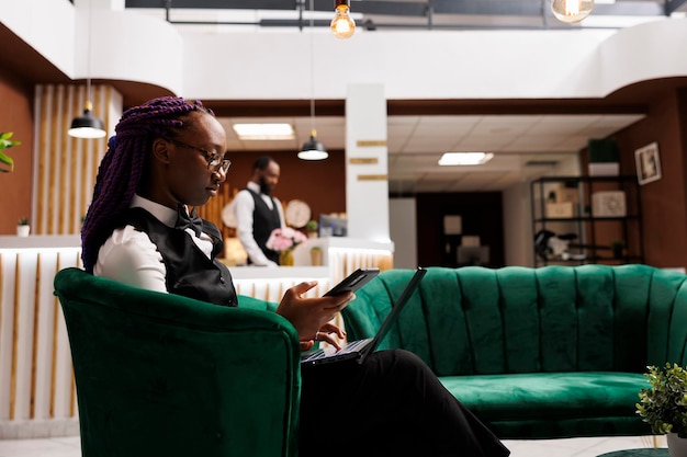 Jonge Afro-Amerikaanse hotelmanager zit in de receptie met een smartphone en laptop, beheert het budget en bestelt benodigdheden. Technologie in de horeca