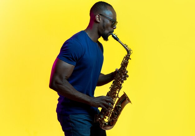 Jonge Afro-Amerikaanse jazzmuzikant die saxofoon speelt op gele studioachtergrond in trendy neonlicht. Concept van muziek, hobby. Vrolijke man improviseren. Kleurrijk portret van kunstenaar.