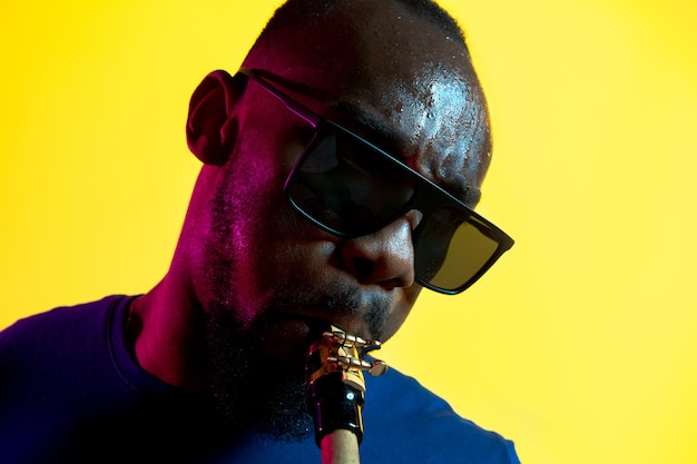 Jonge Afro-Amerikaanse jazzmuzikant saxofoon spelen op gele achtergrond in trendy neonlicht.