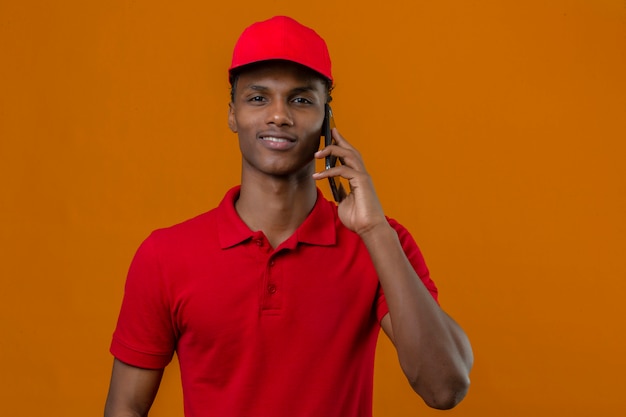 Jonge Afro-Amerikaanse levering man met rode polo shirt en pet spreken door smartphone op zoek zeker over geïsoleerde oranje
