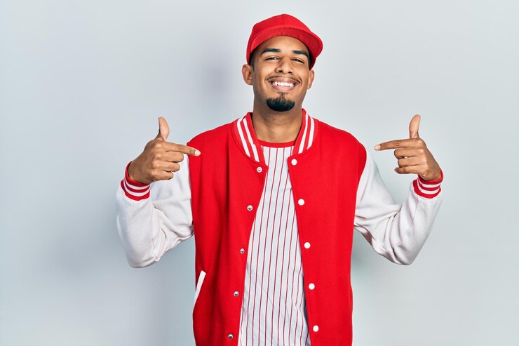 Jonge afro-amerikaanse man in honkbaluniform die er zelfverzekerd uitziet met een glimlach op het gezicht, zichzelf wijzend met vingers trots en gelukkig