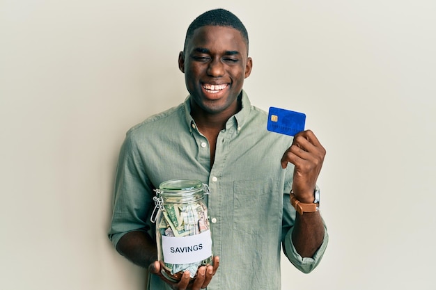 Jonge afro-amerikaanse man met creditcard en pot met dollars glimlachend en hard hardop lachend omdat grappige gekke grap.